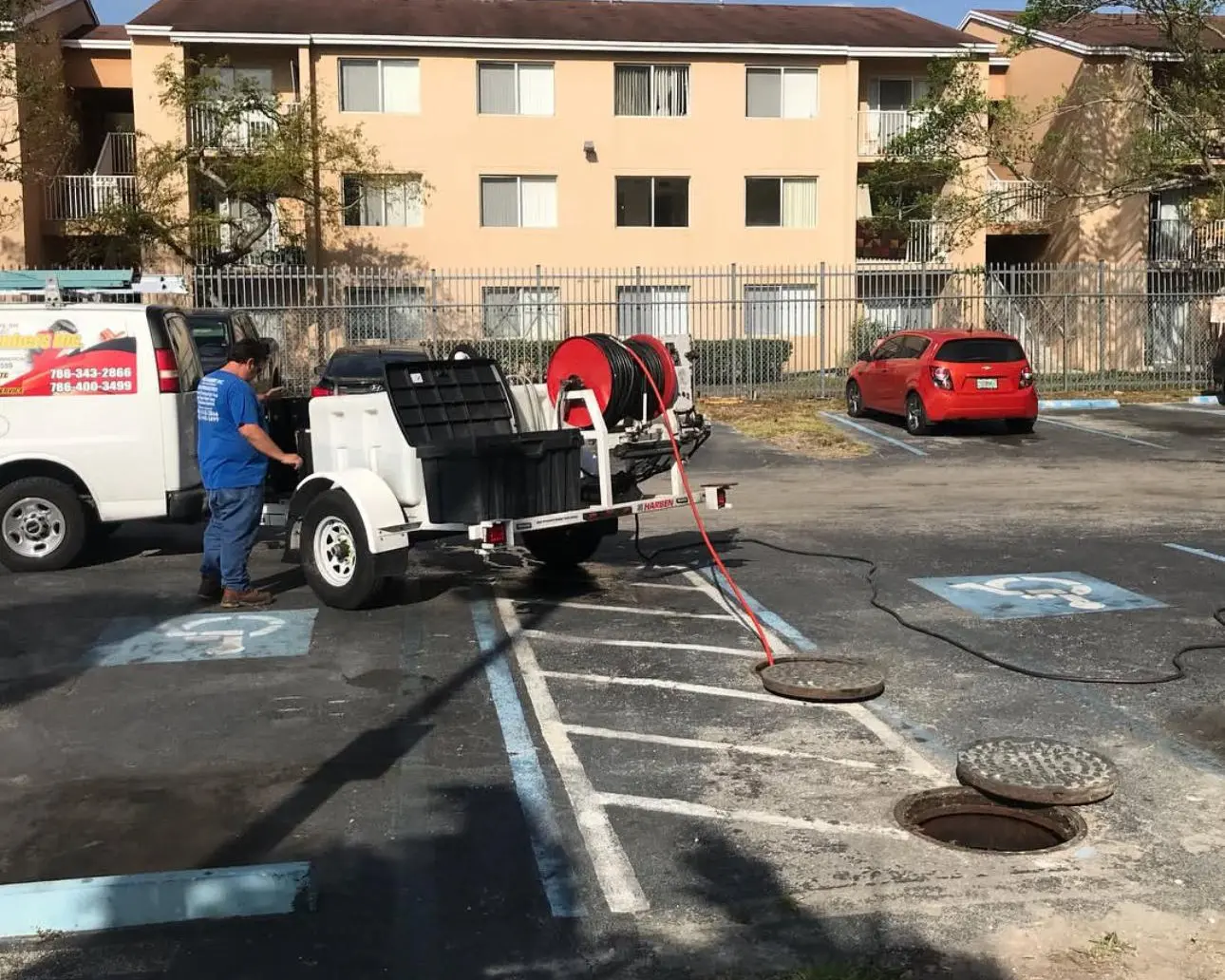 A D’Plumbers Inc. technician operates a high-pressure sewer jetting machine in a residential parking lot, clearing sewer lines through open manholes using red jetting hoses.
