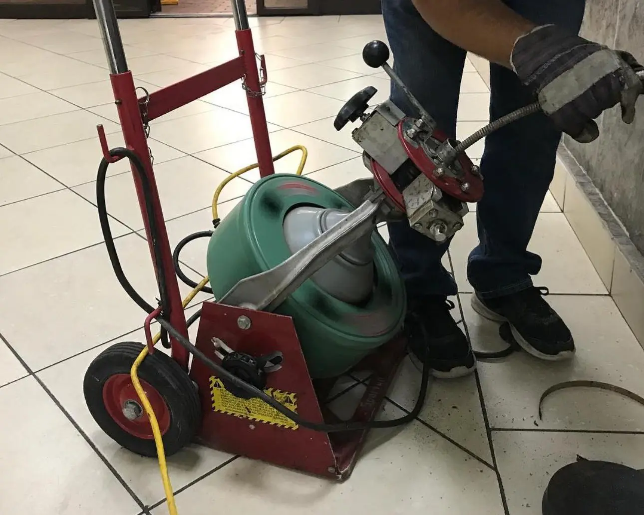 Close-up of a plumber operating a mechanical drain snake machine to clear a sewer line clog in a tiled indoor area, showing the flexible cable being fed into the drain.
